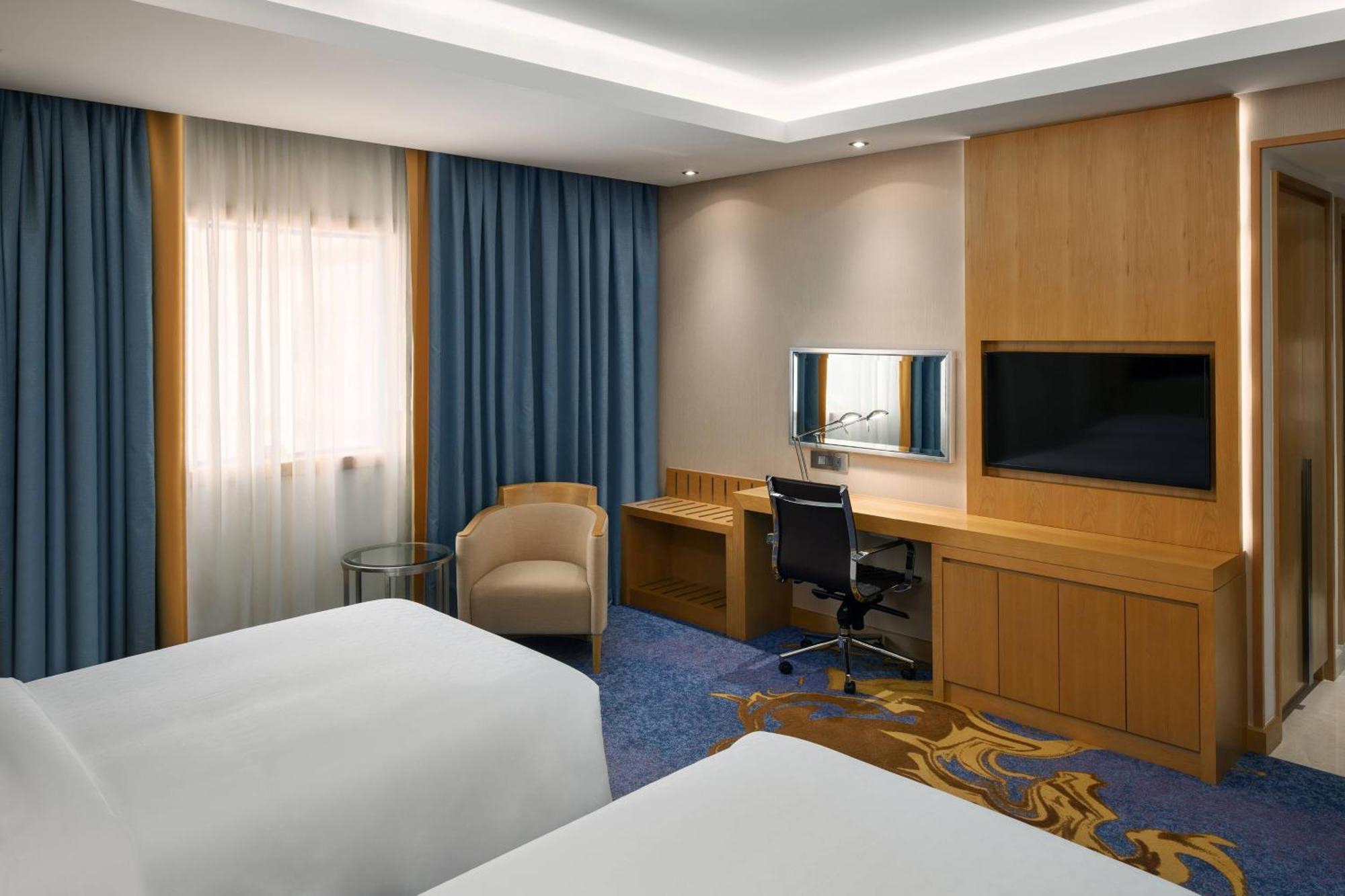 Sheraton Makkah Jabal Al Kaaba Hotel Mekka Exterior foto The photo shows a hotel room featuring two beds with white linens. There are large windows covered with blue curtains that allow natural light to enter. On one side of the room, there is a wooden workstation with a chair and a mirror, along with a fl