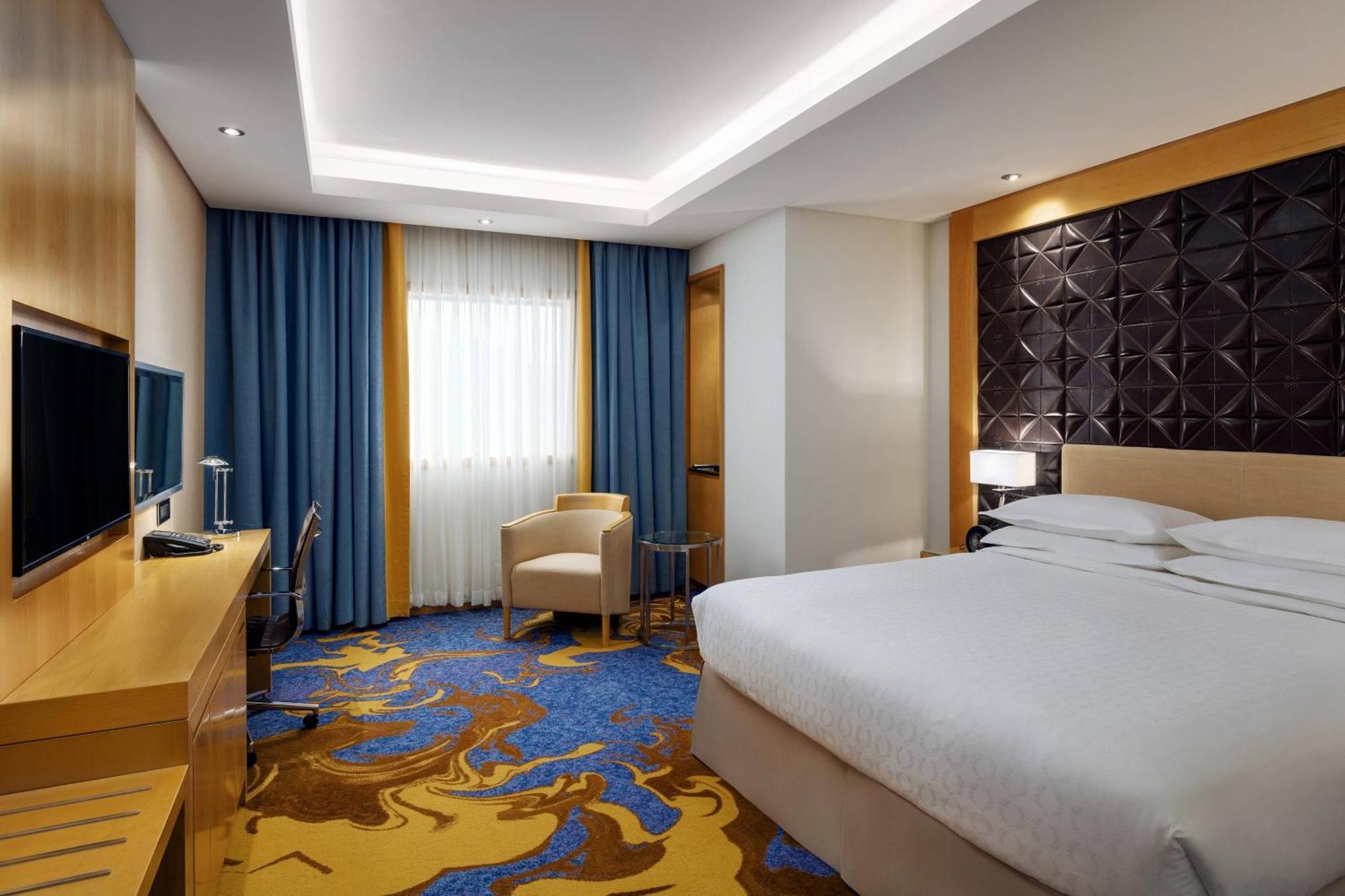 Sheraton Makkah Jabal Al Kaaba Hotel Mekka Exterior foto The photo shows a modern hotel room featuring a comfortable double bed with a light-colored headboard. There's a large window dressed with blue curtains, allowing natural light to enter. The walls are painted in neutral tones, and one wall has a text