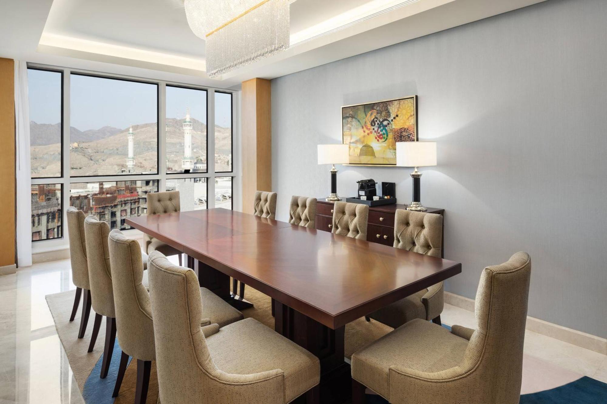 Sheraton Makkah Jabal Al Kaaba Hotel Mekka Exterior foto The photo shows a modern conference room with a large wooden table surrounded by upholstered chairs. The room is well-lit, featuring large windows that offer a view of the outdoors, possibly with mountains in the background. On one wall, there is a p